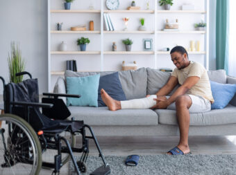 Millennial black man having plastered broken leg, suffering from pain on sofa at home, wheelchair standing nearby